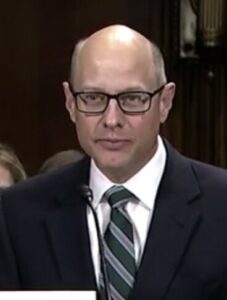 A bald man in a suit and tie speaking into a microphone.