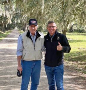Two men standing on a dirt road with spanish moss in the background.