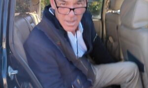 A man in glasses sitting in the back seat of a car.